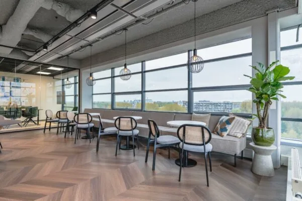 Moderne zithoek in een kantoor aan de Joop Geesinkweg met ronde tafels, designstoelen, grote ramen en uitzicht op een woonwijk.